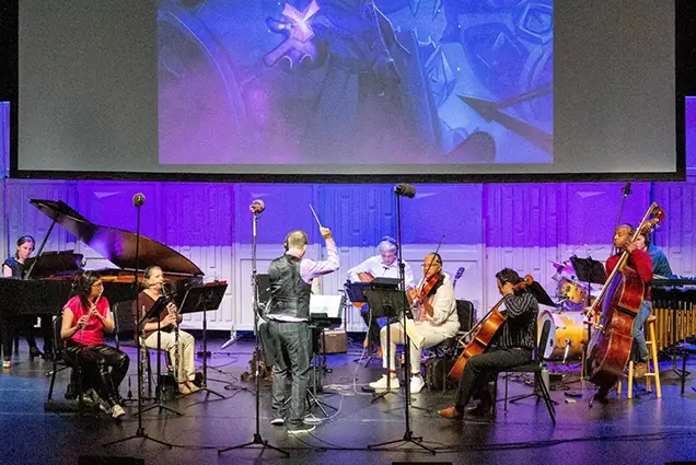 A photo of a small orchestra on stage playing before a projected movie on a screen above them.